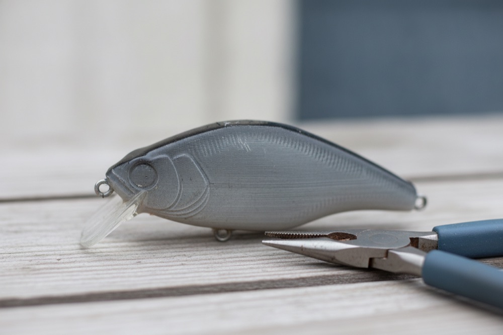 A used 3D-printed crankbait in grey PETG with scuffed clear coat, lying on weathered wood next to fishing pliers